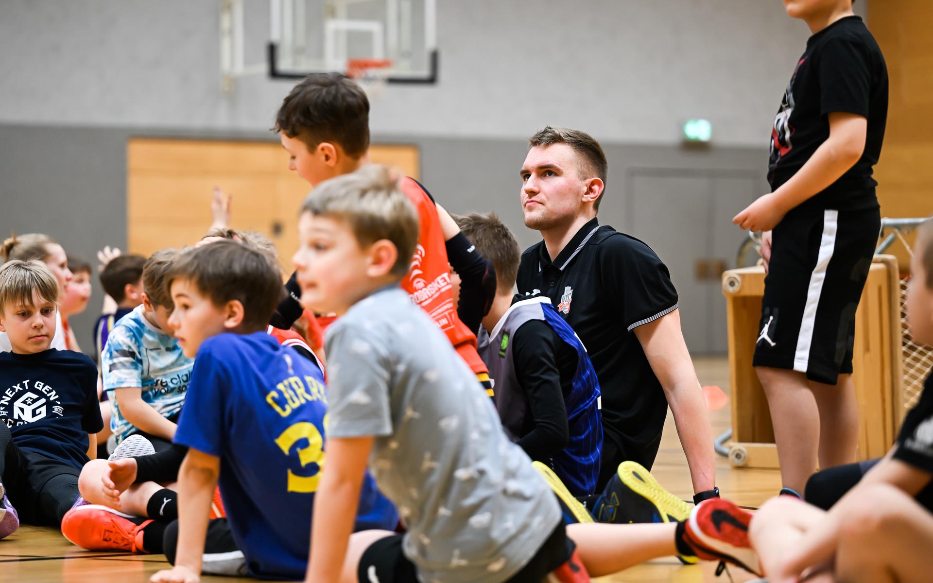 Gruppe von Kindern sitzt auf dem Boden einer Sporthalle, während ein Trainer aufmerksam auf sie schaut.