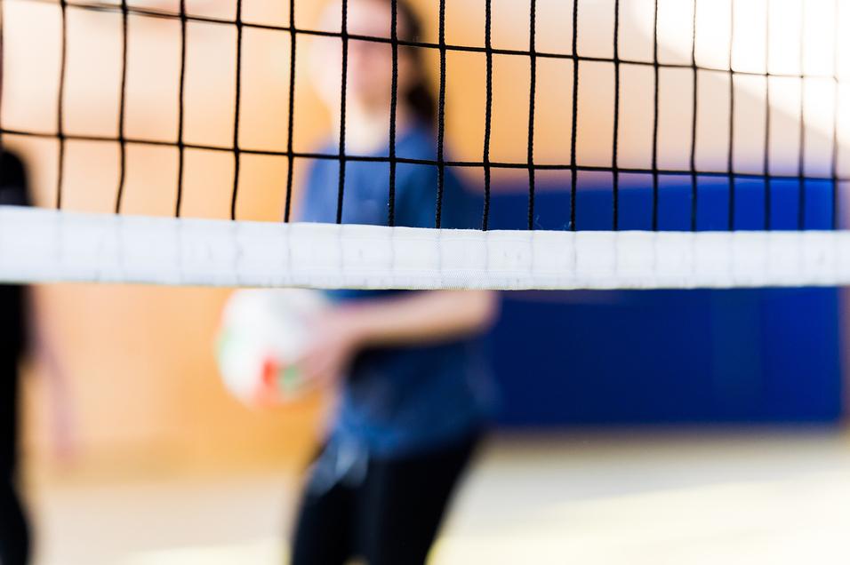 Unscharfe Silhouetten von zwei Personen hinter einem Volleyballnetz in einer Sporthalle. Eine Person hält einen Volleyball.