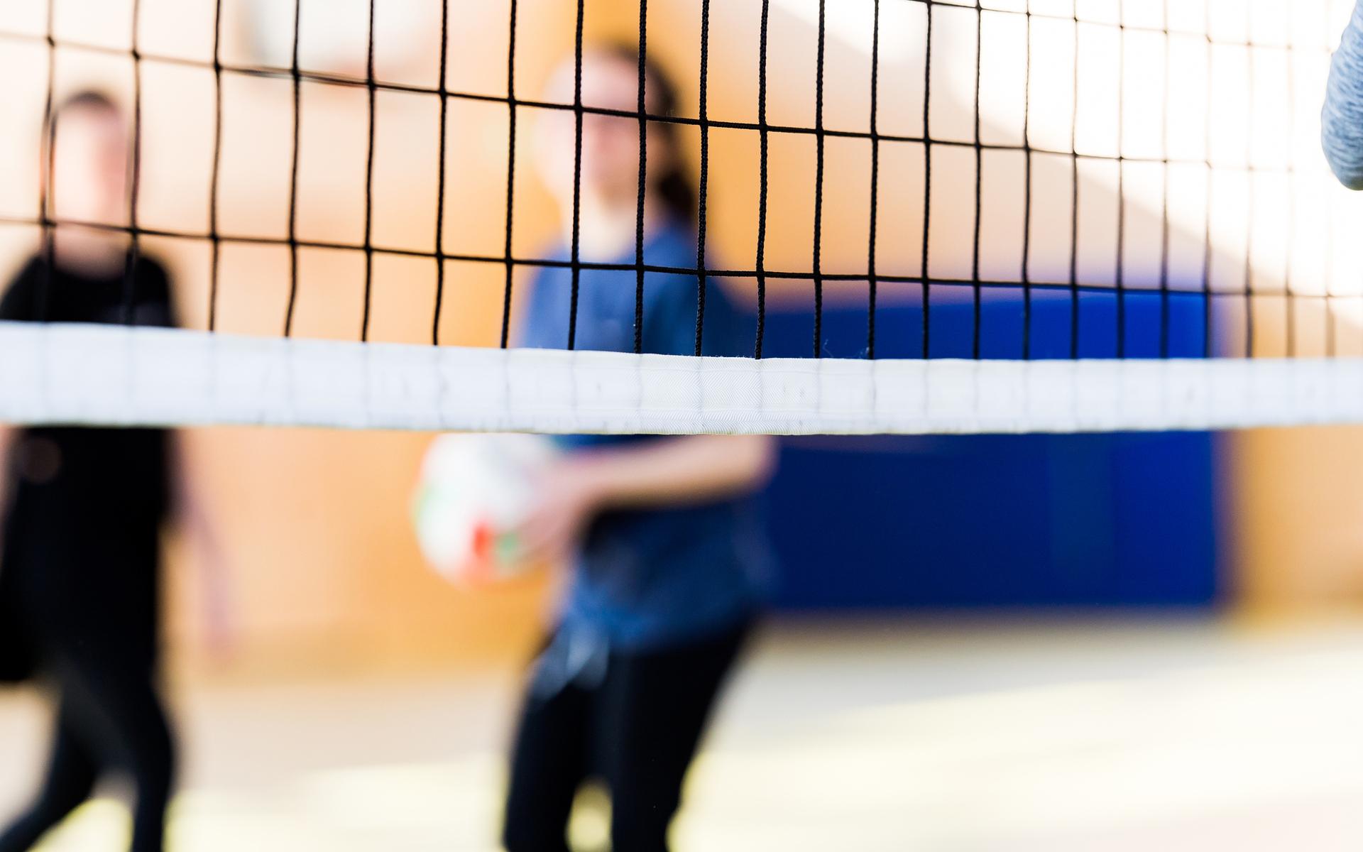 Unscharfe Silhouetten von zwei Personen hinter einem Volleyballnetz in einer Sporthalle. Eine Person hält einen Volleyball.